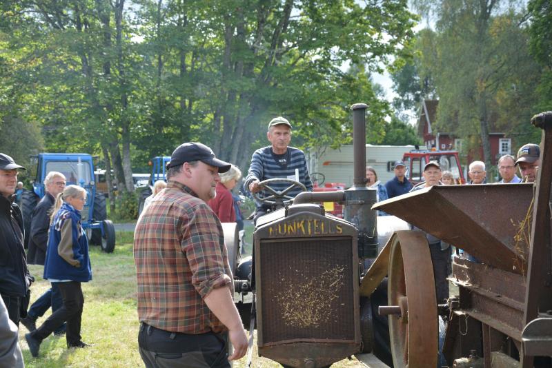 En grupp människor samlas utomhus runt en man som kör en gammal Lanz Bulldog-traktor, med träd och byggnader synliga i bakgrunden.