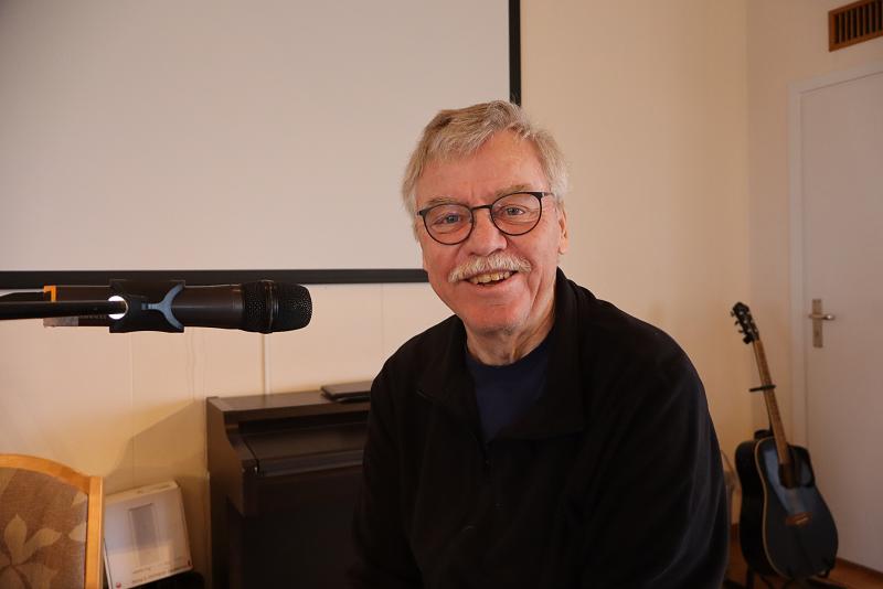 Leende äldre man med glasögon sitter vid en mikrofon, med gitarr och piano i bakgrunden.
