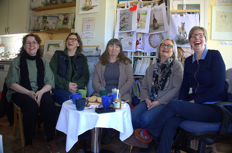 Fem kvinnor sitter runt ett litet bord med drinkar och snacks, ler och skrattar i ett rum inrett med djurkonstverk och bokhyllor.