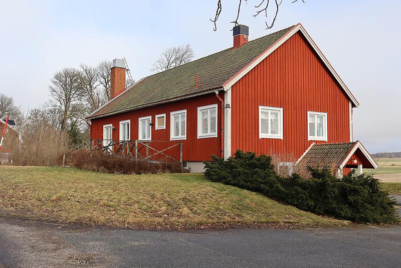 Ett rött trähus med grönt tegeltak ligger på en gräsbevuxen tomt nära en asfalterad väg, omgivet av träd och buskar under en molnig himmel.