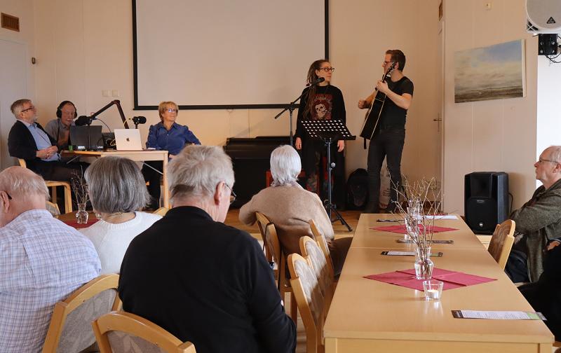 En liten publik lyssnar på två musiker som uppträder på scenen medan tre personer sitter vid ett bord med mikrofoner och bärbara datorer i ett mötesrum.