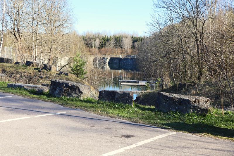En asfalterad väg med stora stenar i kanten, träd utan löv och en lugn flod med en stenkonstruktion och en brygga i bakgrunden.