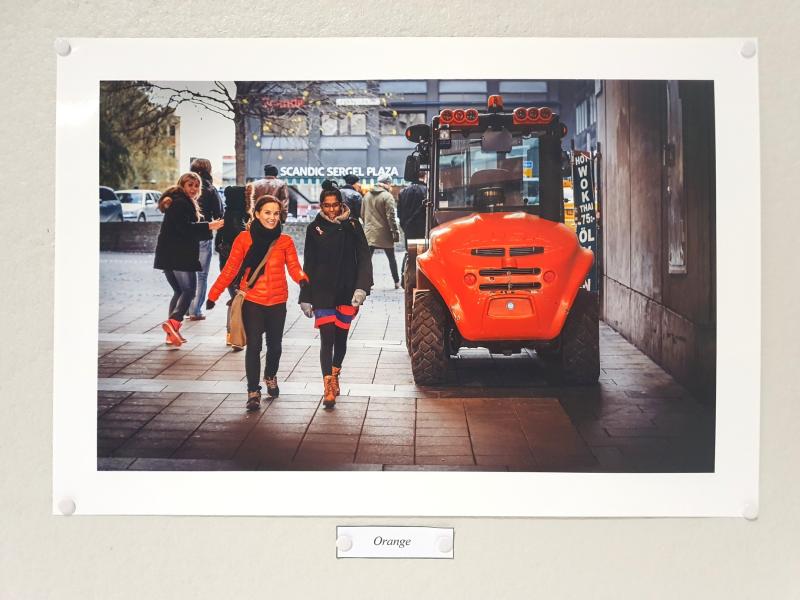 Två kvinnor går på en trottoar i en stad förbi ett orange byggfordon; andra fotgängare och byggnader syns i bakgrunden.