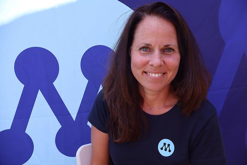 En kvinna med långt brunt hår sitter framför en blå bakgrund med en stiliserad ”M”-logotyp. Hon bär en matchande t-shirt och ler mot kameran.