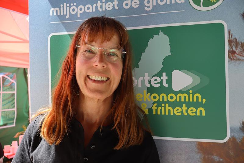 En kvinna med axellångt brunt hår och glasögon som ler framför en skylt med texten ”Miljöpartiet de gröna” vid ett utomhusarrangemang.