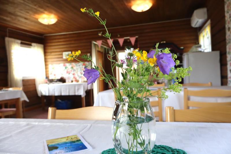 En glasvas med olika vildblommor står på en vit duk i en mysig matsal med träinredning och enkel inredning.