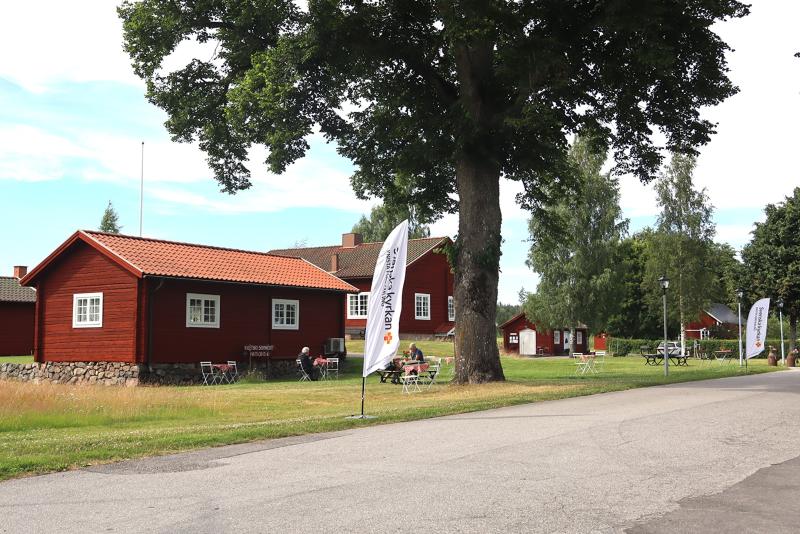 Röda träbyggnader med vita lister står intill en väg, omgivna av träd och gräs. Vita flaggor samt utomhusbord och stolar syns. Himlen är delvis molnig.