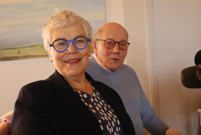 En äldre kvinna och en man med glasögon sitter inomhus och ler mot kameran. En landskapsmålning hänger på väggen bakom dem.