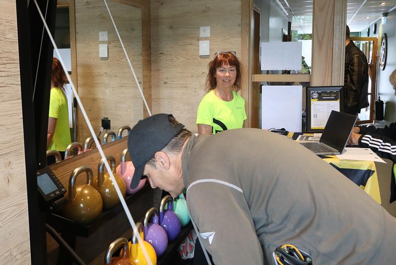 En man inspekterar färgglada kettlebells på ett ställ medan en kvinna i gul skjorta står bakom en disk och ler.