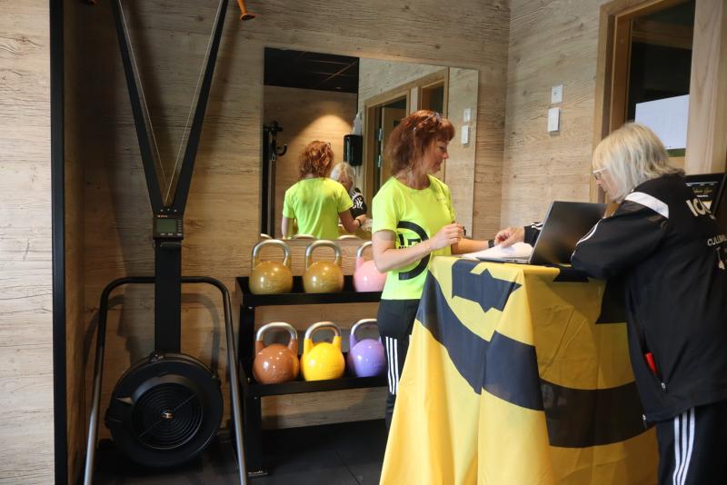 Två kvinnor står vid en receptionsdisk på ett gym som är täckt av en gul och svart duk; kettlebells och en roddmaskin syns i bakgrunden.