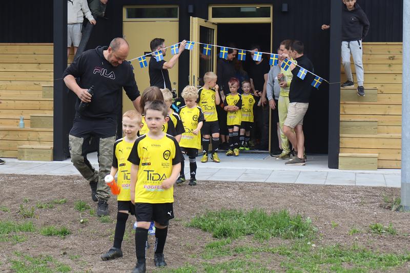 Unga pojkar i gula fotbollsdräkter går ut ur en byggnad, ledda av en vuxen. Andra tittar på och håller ett snöre med små svenska flaggor ovanför entrén.