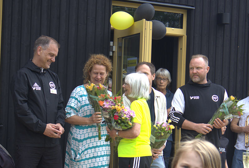 En grupp människor står utanför en byggnad. Flera personer håller i blombuketter och vid dörröppningen står gula och svarta ballonger.