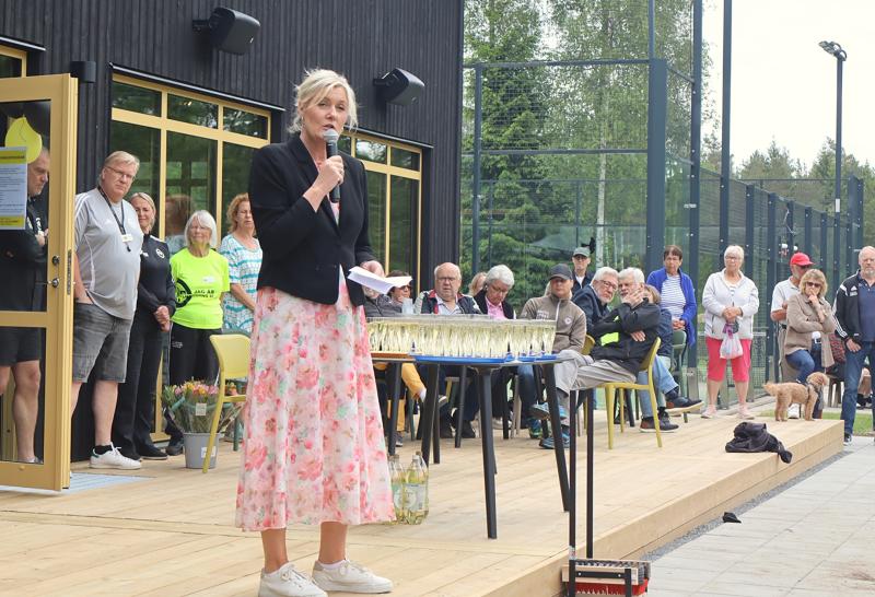 En kvinna i blommig kjol talar i en mikrofon på ett trädäck med sittande och stående människor, drinkar på ett bord och träd i bakgrunden.