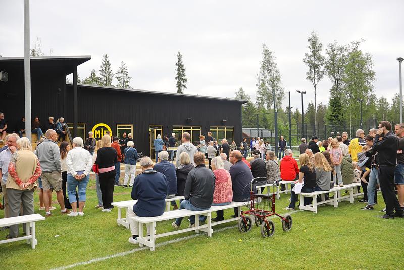 En grupp människor är samlade utanför en svart byggnad, några sittande på bänkar och andra stående, på en gräsyta omgiven av träd.