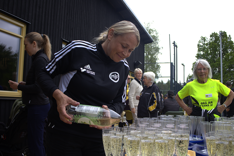 En kvinna i sporttröja häller upp mousserande dryck i plastmuggar utomhus, med människor samlade runt ett bord.