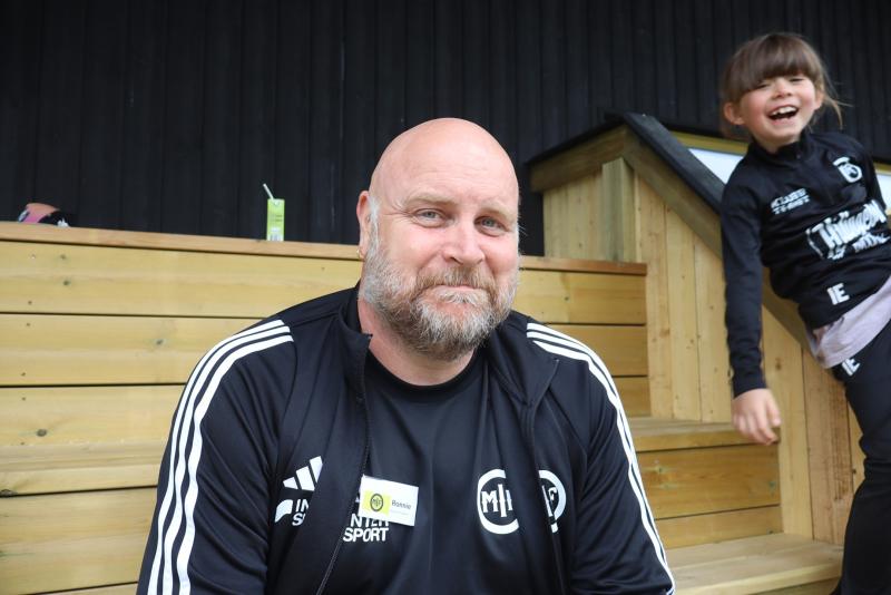 En man i svart sportjacka sitter på en läktare av trä och ler mot kameran, medan ett barn i sportkläder skrattar i bakgrunden.