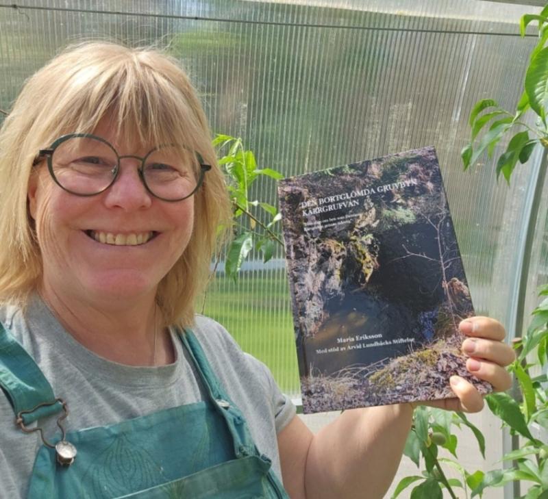 En leende person med glasögon och blont hår håller upp en bok med ett omslag med naturtema när han står i ett växthus.