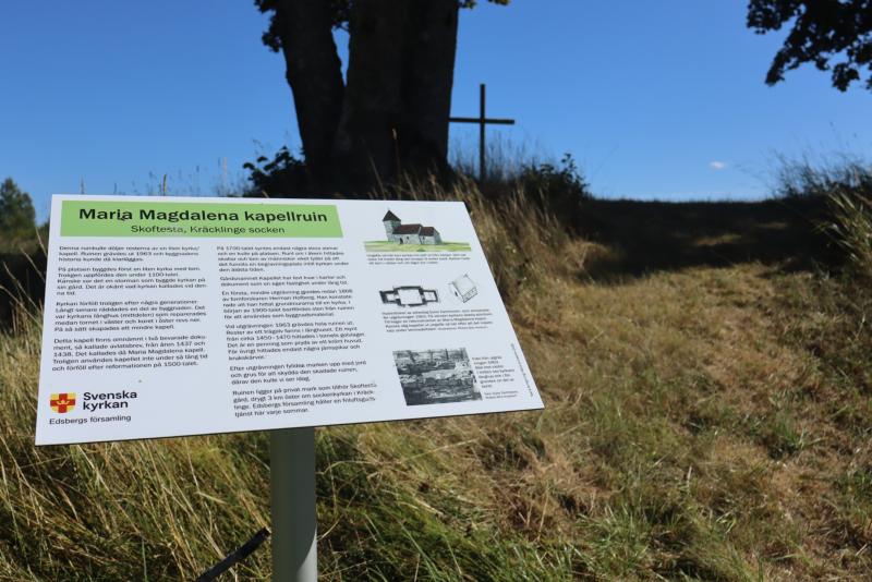 En informationsskylt om ruinerna av Maria Magdalena-kapellet står vid en gräsbevuxen stig som leder uppför backen till ett stort träkors under ett träd, med en klarblå himmel i bakgrunden.