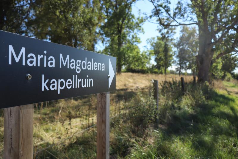 En svart skylt med texten ”Maria Magdalena kapellruin” och en pil, placerad nära en gräsbevuxen äng och träd en solig dag.