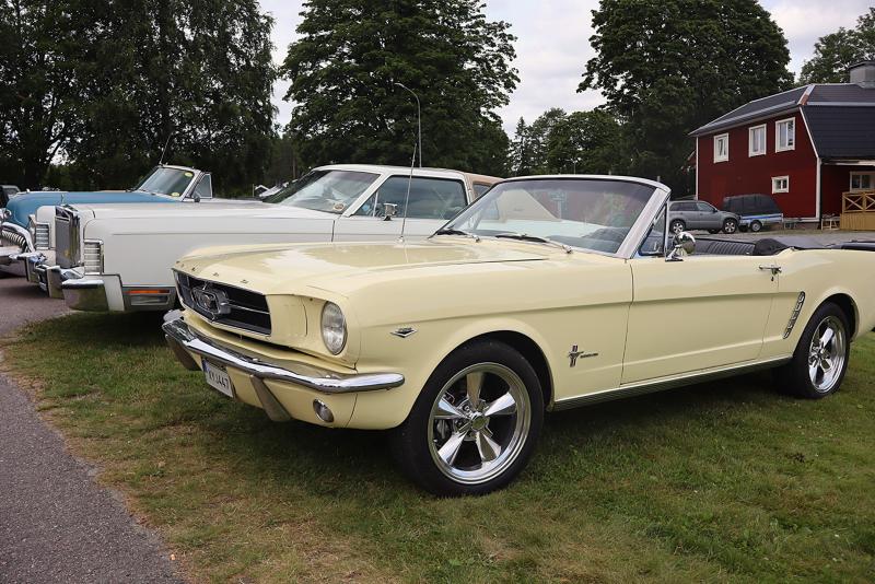 En gul Ford Mustang-cabriolet i vintage-stil står parkerad på gräset bredvid två klassiska bilar, med träd och ett rött hus i bakgrunden.