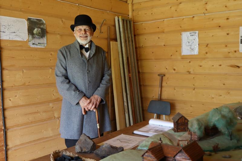 En äldre man i grå rock och svart hatt står i ett trärum bredvid ett bord med kartor och byggmodeller i miniatyr.