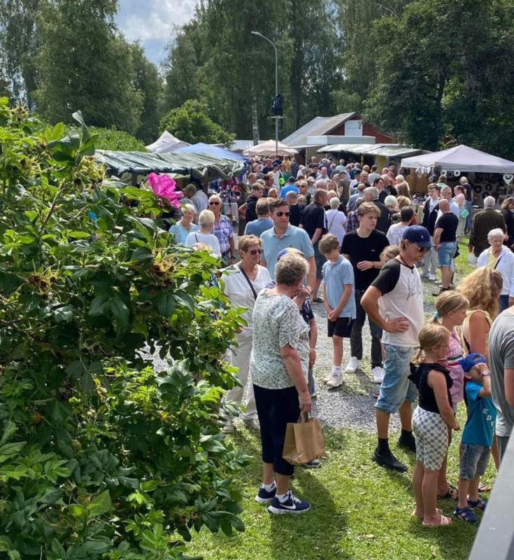 En stor folkmassa promenerar och samlas på en utomhusmarknad eller marknad en solig dag, omgiven av träd och försäljartält, med grönska i förgrunden.