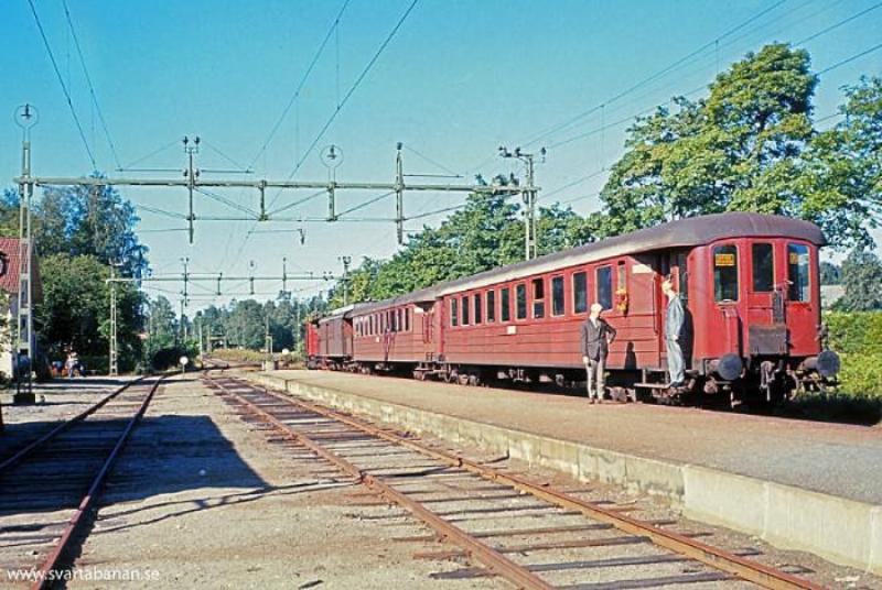 Ett rött passagerartåg har stannat på en perrong på en lantlig station. Två personer står vid sidan av tåget. I bakgrunden syns kraftledningar och träd.