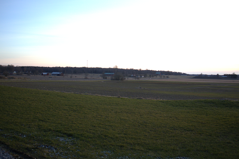 Ett stort, öppet fält med grönt gräs och lantbruksbyggnader i fjärran under en klar himmel.