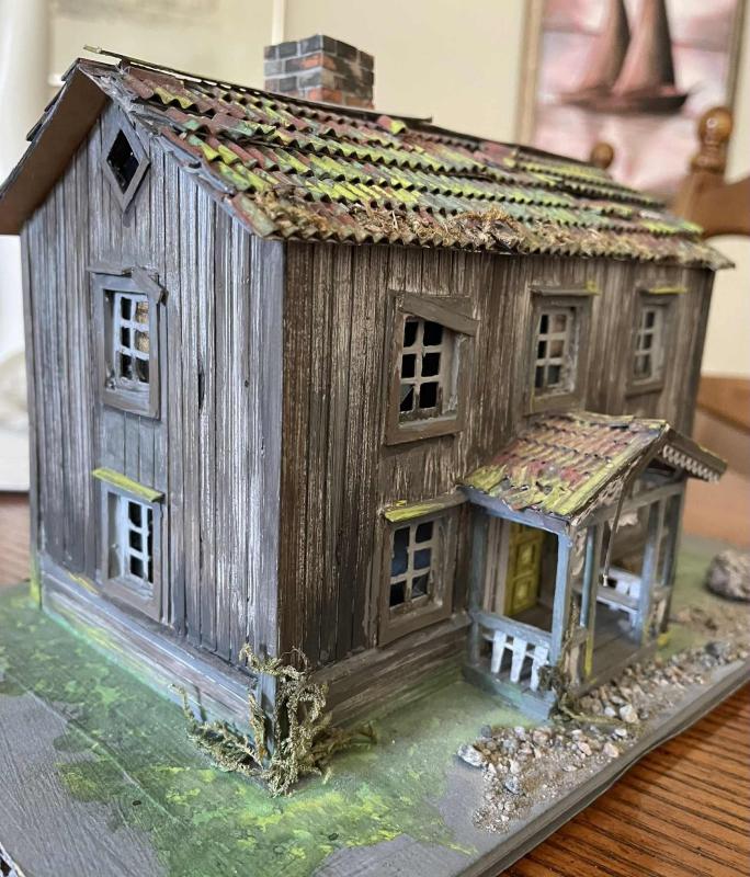 Detaljerad modell av ett väderbitet trähus med flagnande färg, mossbelupet tak och liten veranda, på en texturerad bas.