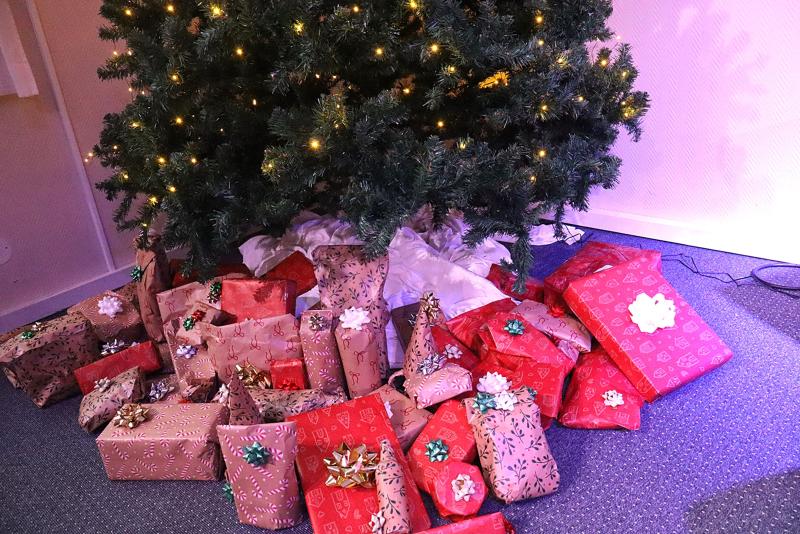 En grupp inslagna julklappar med rosetter är placerade under en dekorerad julgran med lampor, på ett golv med heltäckningsmatta.