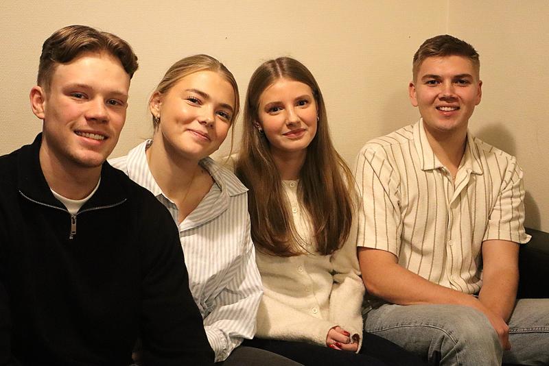 Fyra unga vuxna sitter sida vid sida i en soffa, vänder sig mot kameran och ler i ett varmt upplyst rum med en enfärgad väggbakgrund.