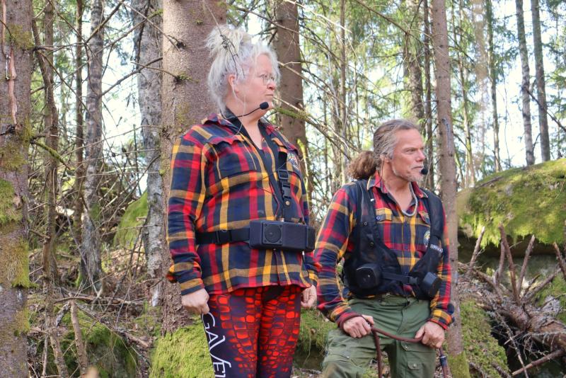 Två personer i rutiga skjortor står i en skog, den ena bär rödmönstrade leggings och headset, den andra håller i en pinne. Båda har friluftsutrustning fäst på sina skjortor.