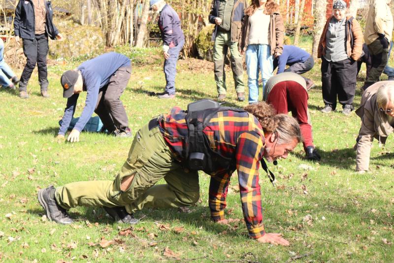 En grupp människor utomhus på en gräsmatta utför en kryp- eller stretchövning, några av dem bär friluftskläder och ryggsäckar.