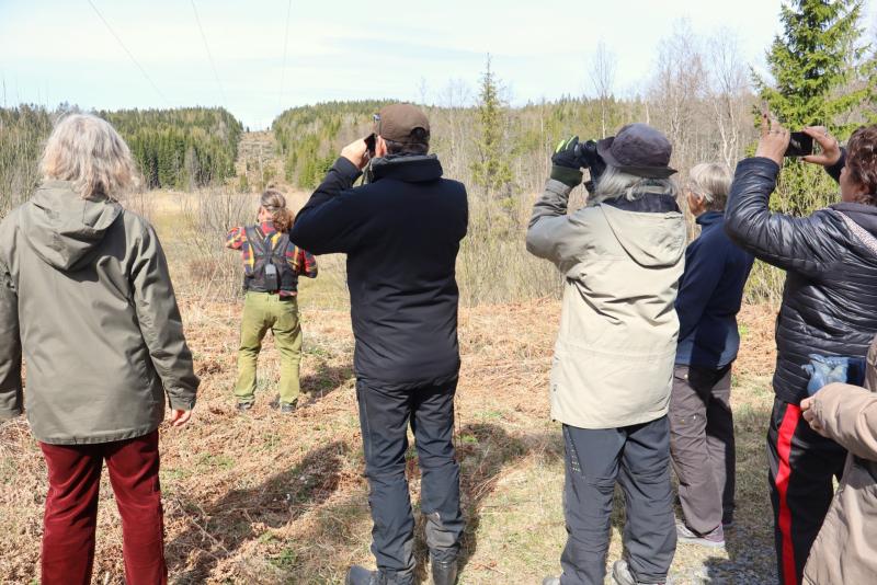 En grupp människor står utomhus i ett naturområde, några använder kikare för att observera landskapet framför sig.