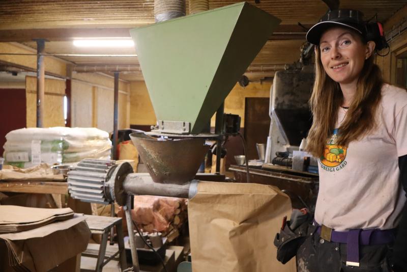 En kvinna med keps och arbetskläder står bredvid en grön industrimaskin i en verkstad, med bruna papperspåsar och förnödenheter i bakgrunden.