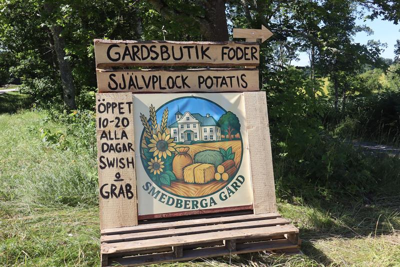 Träskylt för Smedberga Gård med information om gårdsbutik, djurfoder och självplockning av potatis. Öppet dagligen kl. 10–20. Betalning med Swish går att använda.