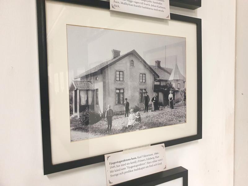 Inramat svartvitt fotografi av en familj som poserar utanför ett tvåvåningshus, placerat på en vit vägg med bildtexter under ramen.