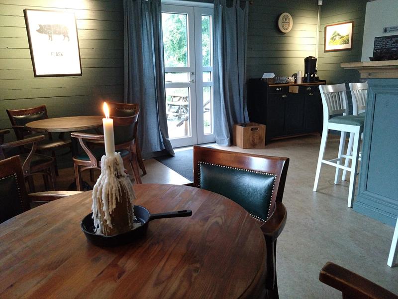 Ett tänt ljus i en stekpanna står på ett träbord i ett mysigt café med trästolar, runda bord och stora fönster i bakgrunden.