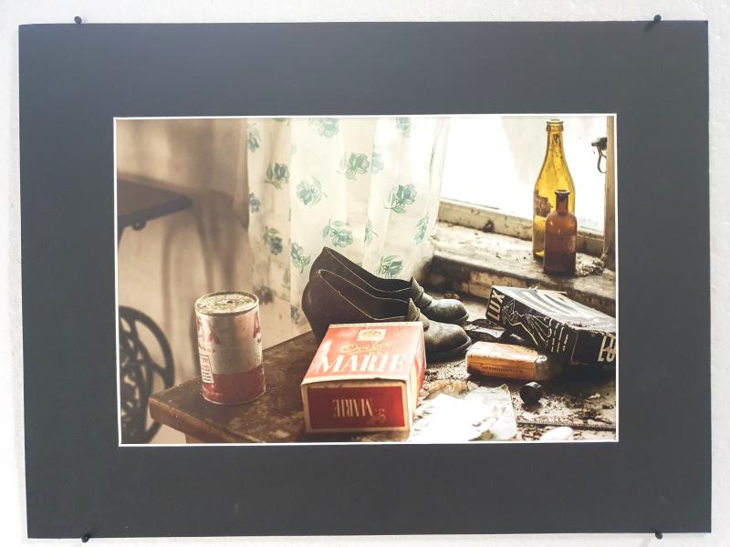 Ett fotografi av ett gammalt bord med en Mariboro cigarettask, slitna skor, konserver, flaskor och diverse skräp framför ett fönster med blommiga gardiner.