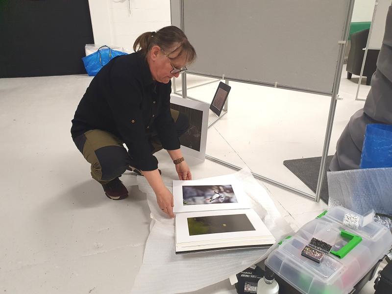 En person står på knä på golvet och arrangerar fotografiska utskrifter på förpackningsmaterial nära en låda med material i ett galleri eller en studio.