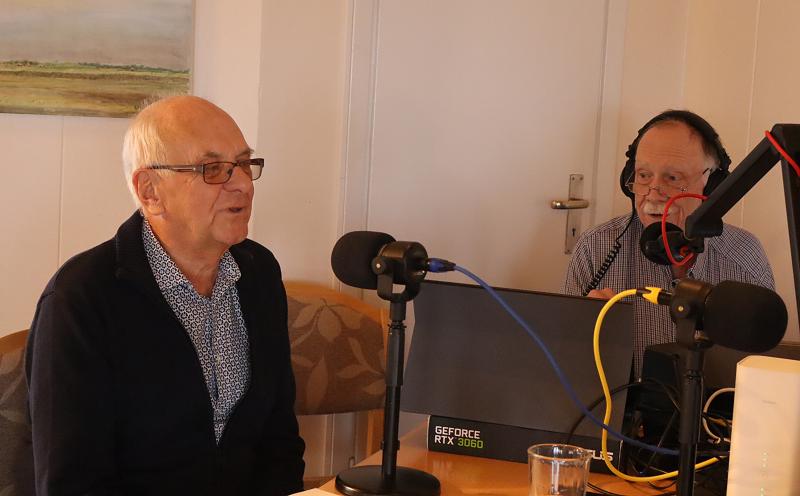 Två äldre män sitter vid ett bord framför mikrofoner och inspelningsutrustning och ser ut att delta i en podcast eller radiointervju.