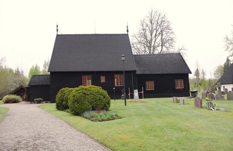En svart träkyrkobyggnad med rödkarmade fönster, omgiven av grönt gräs, buskar, gravstenar och en grusgång under en mulen himmel.