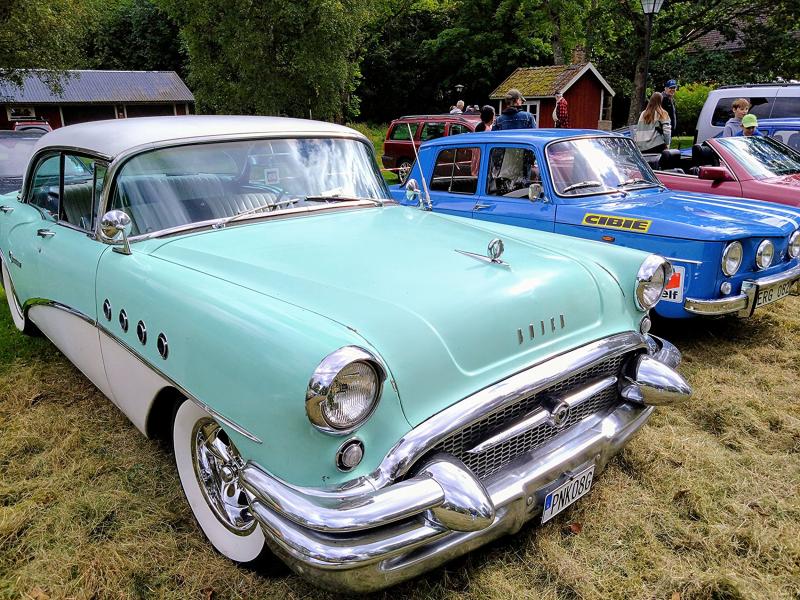 En ljusgrön vintage Buick och en blå rallybil står parkerade på gräset vid en klassisk bilutställning, med människor som står och tittar på fordonen i bakgrunden.