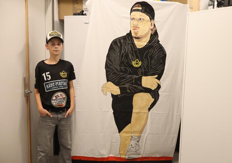 En pojke i svart skjorta och keps står bredvid en stor tygbanderoll som föreställer en man i huvtröja och keps, knäböjande med en hand på sitt knä.