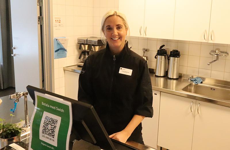 En kvinna i svart jacka står bakom en disk i ett kök med kaffeautomater, diskbänk och en betalskylt med en QR-kod i förgrunden.