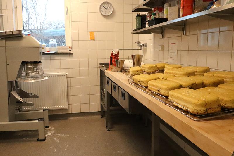 Rektangulära sockerkakor som svalnar på galler i ett storkök med kaklade väggar, en mixer och olika baktillbehör på hyllor.