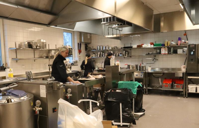 Tre personer arbetar i ett storkök med utrustning i rostfritt stål, hyllor med varor, en diskbänk och olika rengöringsmedel synliga.