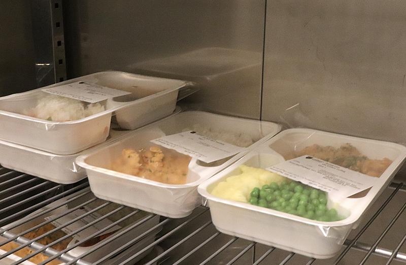 Flera färdigförpackade måltider i vita brickor med genomskinliga plastlock och etiketter är placerade på metallhyllor i ett kylskåp eller en förvaringsenhet.