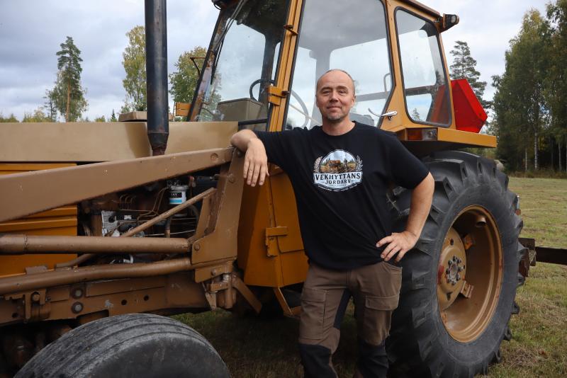 En person står bredvid en brun traktor på en gräsbevuxen åker med träd i bakgrunden och poserar med ena handen på traktorn och den andra på höften.