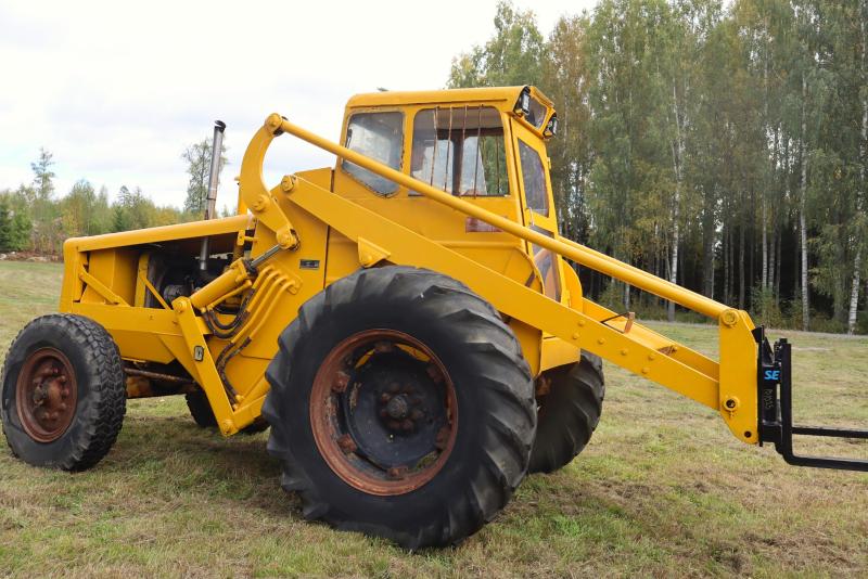 En gul traktor med stora svarta däck står parkerad på gräset, med ett skogsområde i bakgrunden.